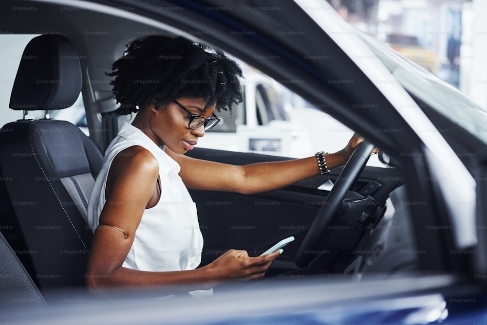 Using phone. Young african american woman sits inside of new modern car.