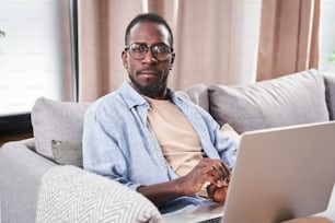 다인종 수염을 기른 남자의 초상화는 노트북으로 작업하고 거실에 앉아 웃고 카메라를 바라보고 있다. 소파에서 노트북으로 일하는 잘생긴 청년. 기술 개념입니다. 스톡 사진