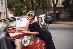 Mulher loira de óculos escuros e de vestido preto apoiada no velho carro clássico vintage.