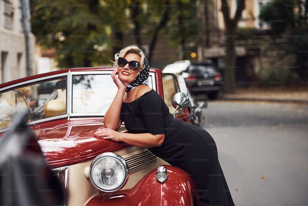 Blonde woman in sunglasses and in black dress leaning on old vintage classic car.