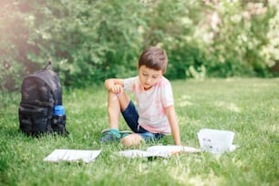 Young Caucasian school boy sitting in park outdoor doing school homework. Child kid writing in notebook with pencil outside. Self education learning studying. Early development for children.
