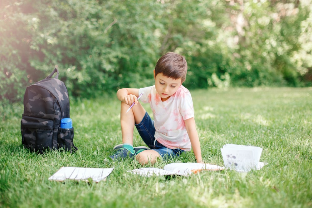 Young Caucasian school boy sitting in park outdoor doing school homework. Child kid writing in notebook with pencil outside. Self education learning studying. Early development for children.