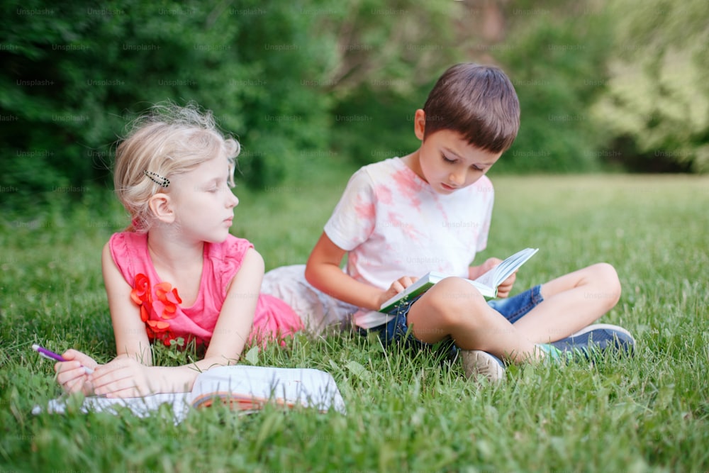 Young Caucasian school girl and boy friends doing school homework in park outdoor. Children kids reading book and writing with pencil. Children education learning studying together.
