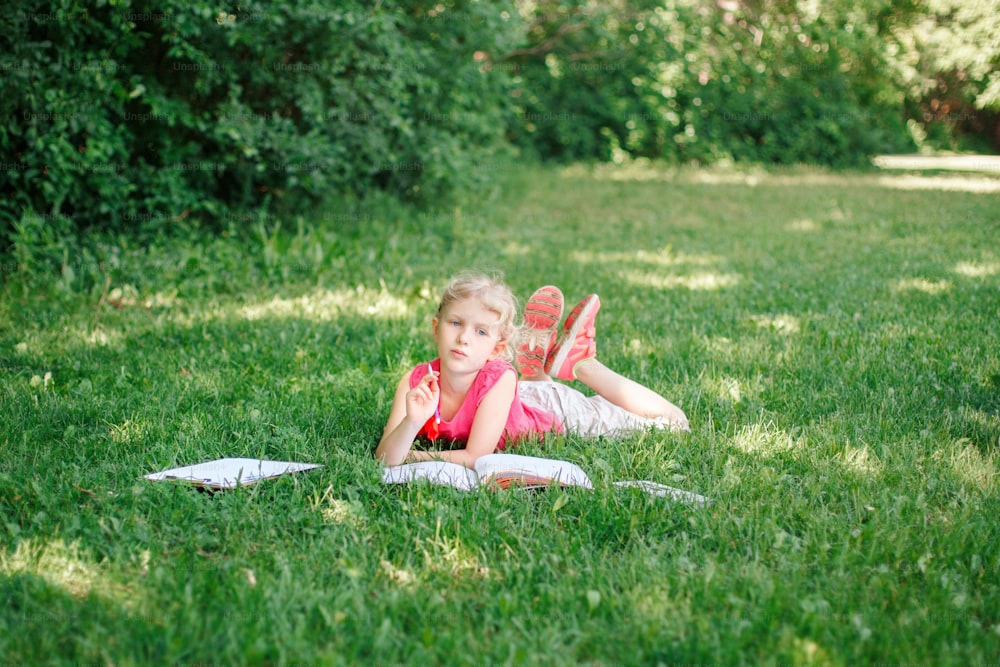 Young Caucasian school girl lying in park outdoor doing school homework. Child kid reading book and writing with pencil. Self education learning studying. Early development for children.