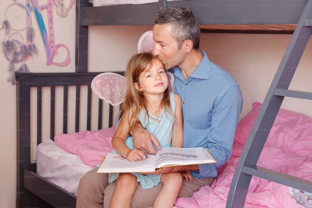 Father and daughters girl at home spending time together. Parent kissing hugging child. Family two people sitting on bed in bedroom reading book. Authentic candid lifestyle. Fathers day holiday.