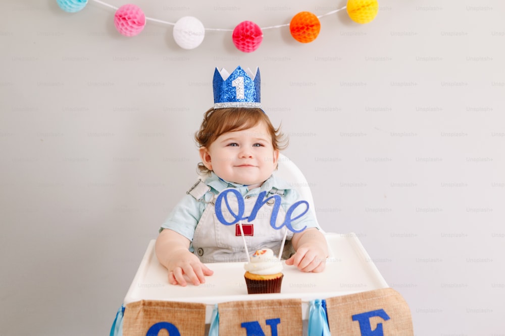 Lindo bebé caucásico sonriente en corona azul celebrando su primer cumpleaños en casa. Niño niño pequeño sentado en silla alta comiendo sabroso postre de magdalenas con la palabra topper Uno. Concepto de feliz cumpleaños.