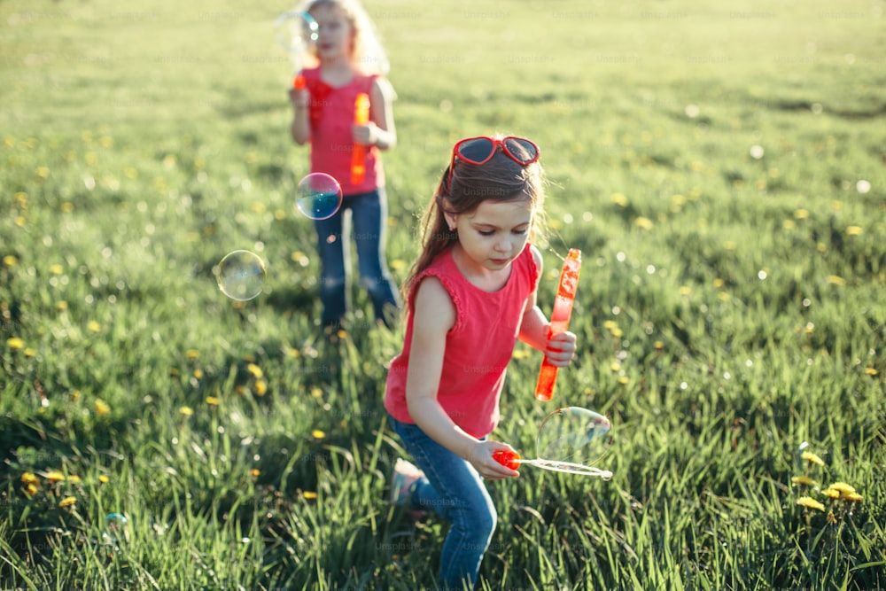 Catch a bubble. Girls friends blowing soap bubbles in park on summer day. Kids having fun outdoor. Authentic happy childhood magic moment. Lifestyle seasonal activity for children.