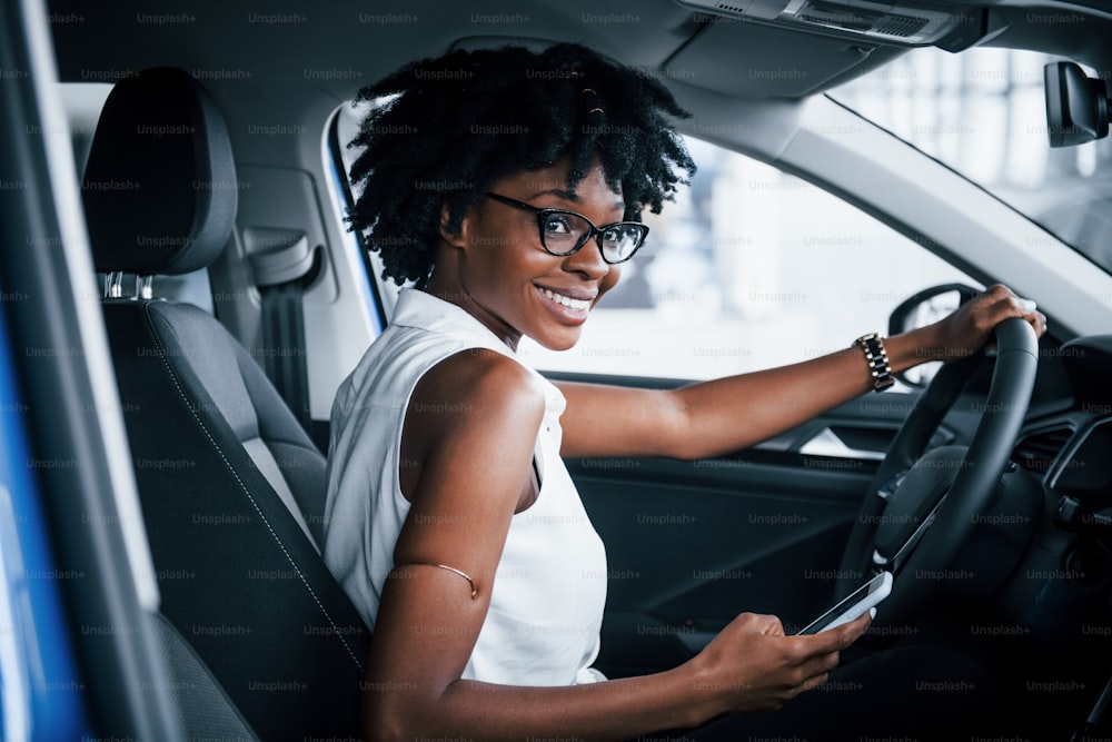 Usando o telefone. Mulher afro-americana jovem senta-se dentro do carro moderno novo.