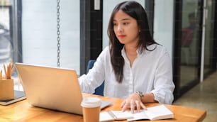 Cropped shot of young beautiful female in white shirt working with laptop and notebook in cafe