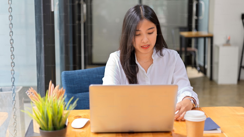 Portrait of young beautiful female in white shirt working with laptop in comfortable office room