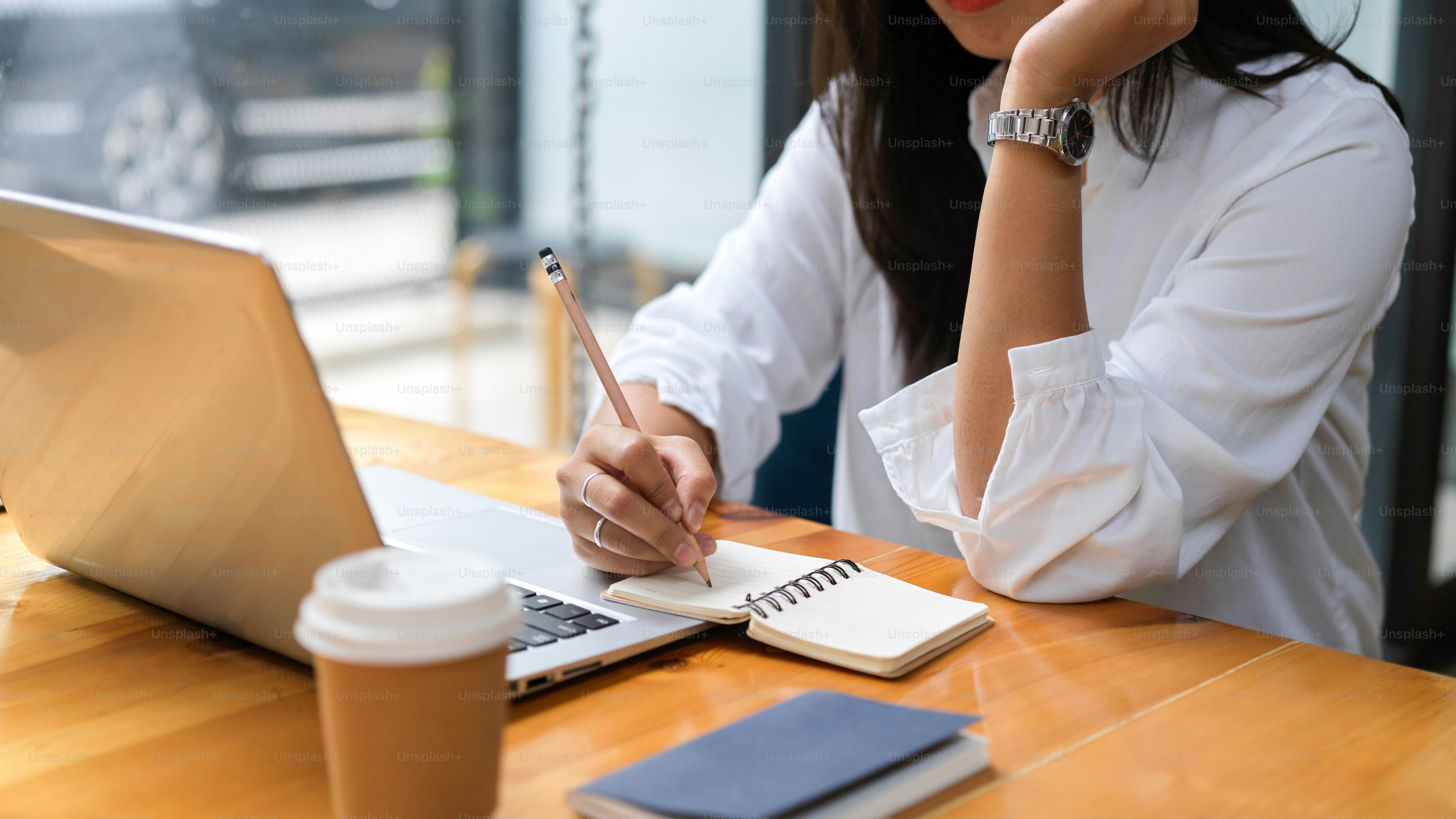 Close up view of female hand with  pencil writing on notebook and using laptop on wooden table in cafe