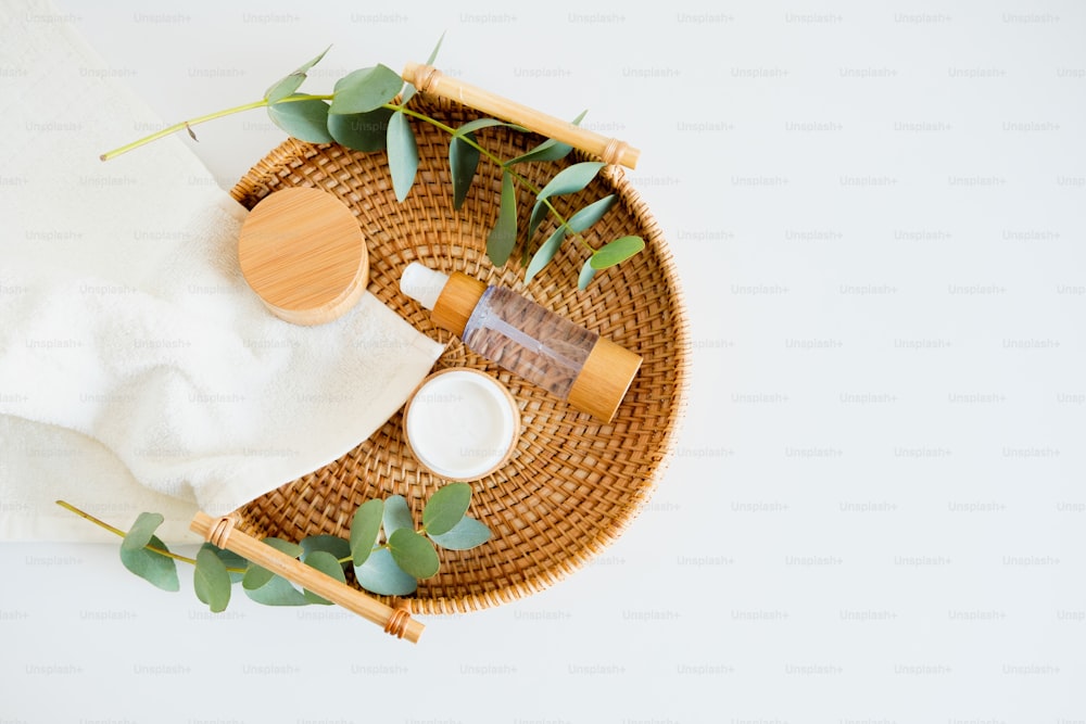 Conjunto de cosméticos orgánicos naturales SPA en bandeja de ratán sobre mesa en baño. Tarro de crema hidratante, loción corporal, hojas de eucalipto, toalla. Plano, vista superior.