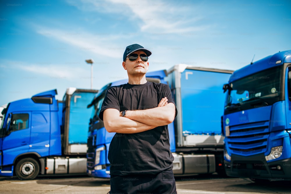 Professional truck driver with hat and sunglasses confidently standing in front of big and modern truck. Bright sunny day. People and transportation concept.