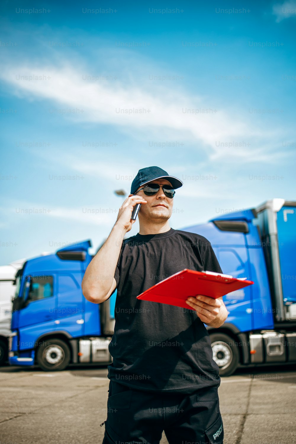 Autista di camion professionista con cappello e occhiali da sole in piedi con sicurezza di fronte a una flotta di camion grande e moderna. Luminosa giornata di sole. Concetto di persone e trasporti.