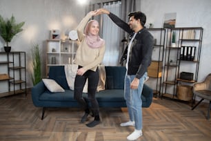 Happy family, young Muslim couple, enjoying being together by dancing indoors, in modern decorated cozy living room. Leisure time, active relaxation.