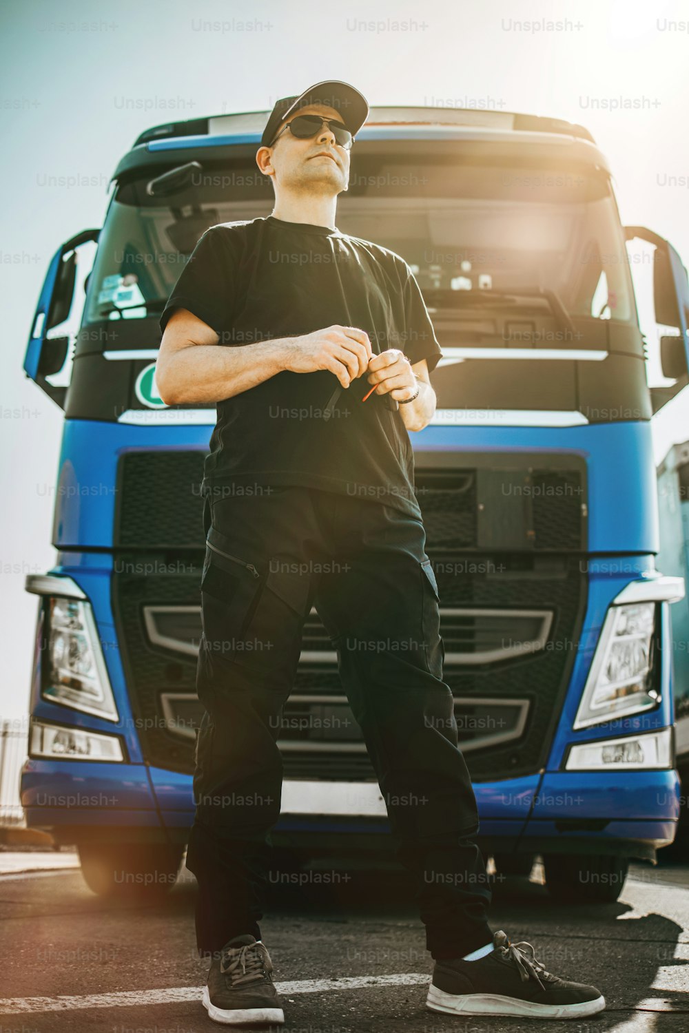 Professional truck driver with hat and sunglasses confidently standing in front of big and modern truck. Bright sunny day. People and transportation concept.