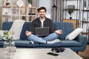 Remote job, relaxation during work, distance online learning. Handsome young Indian man, sitting on sofa at home in lotus position and hands in mudra gesture, relaxing after working on laptop.