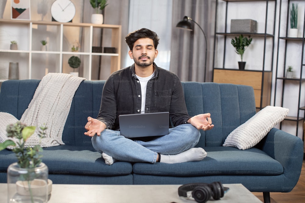 Pleasant likable young arabian indian man, sitting in lotus pose with laptop pc on sofa at living room, and meditating with closed eyes and smile, folding fingers in mudra.
