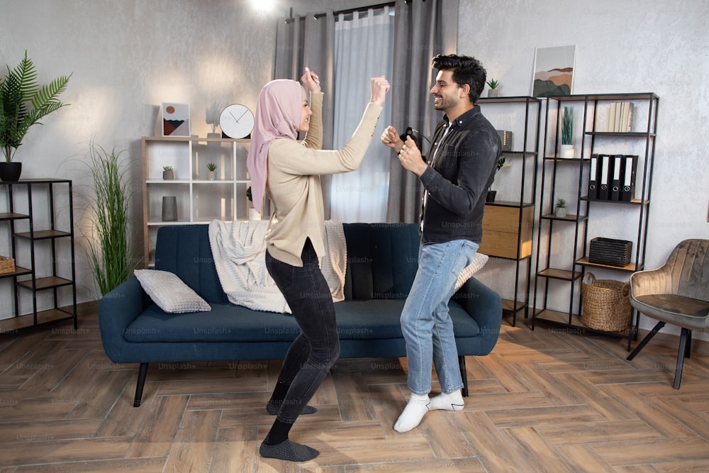 Having fun and nice time at home concept. Bearded young Arabian man dances with his pretty wife in a hijab, spending free evening time together at home living room.