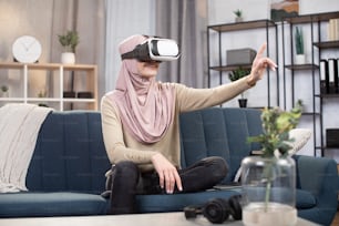 Mujer árabe positiva alegre con hiyab, usando equipos de entretenimiento modernos, divirtiéndose el fin de semana en casa, jugando videojuegos en la sala de estar, usando gafas de realidad virtual