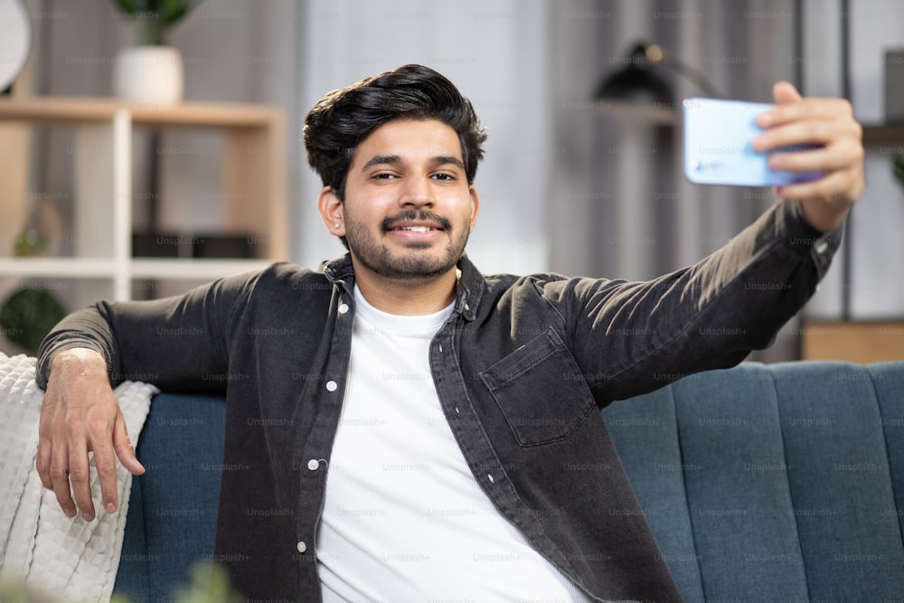 Handsome bearded smiling Indian man, sitting relaxed on comfortable sofa at home and using smartphone for making self photo or having video call. Leisure at home, gadgets concept