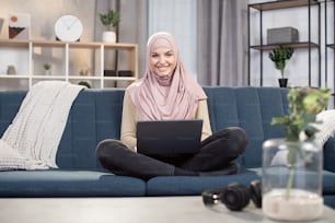 Front view of young muslim woman in hijab, working at home using laptop computer while sitting on sofa, working remotely as a freelancer, studying online, or making shopping.