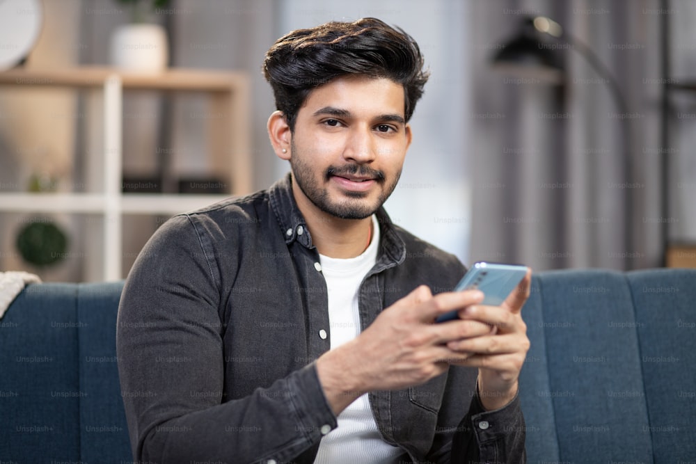 Young smiling Arabian man in casual wear, holding smartphone in hands, sitting on sofa couch, relaxing in living room at home, and looking at camera during leisure time. Close up.