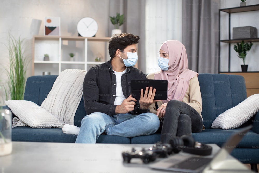 Couple in masks sitting on sofa at home. Young Arabian Muslim couple, spending time during desease and isolation at home, using digital tablet for watching movie or byuing food online