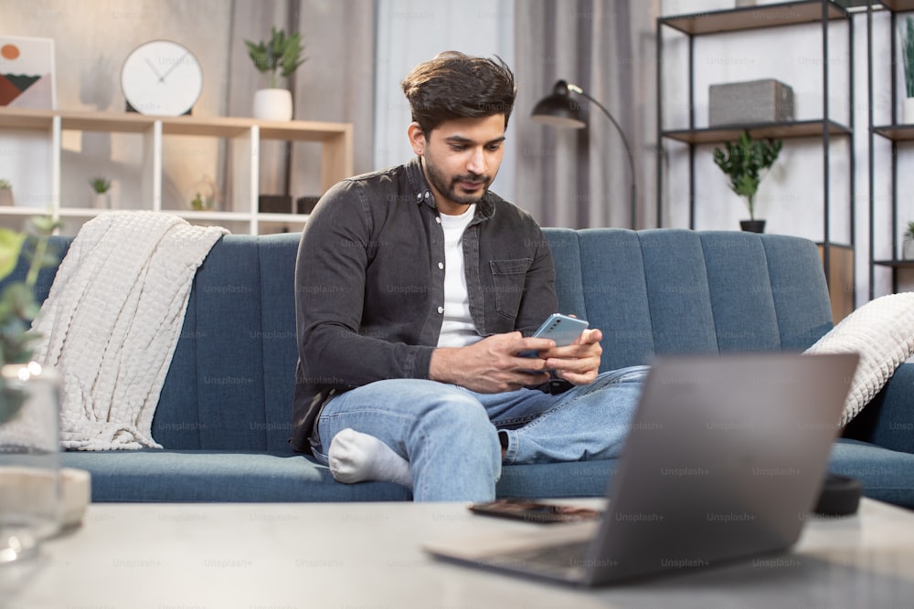 People, home and using gadgets. Young bearded Indian man, sitting on the couch at cozy living room indoors and using smartphone for typing message or serfing social networks.