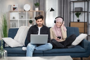 Handsome bearded Arabian guy sitting on blue sofa with laptop together with his muslim girl in headscarf and earphones, using smartphone and listening to music. Young couple enjoying time at home.
