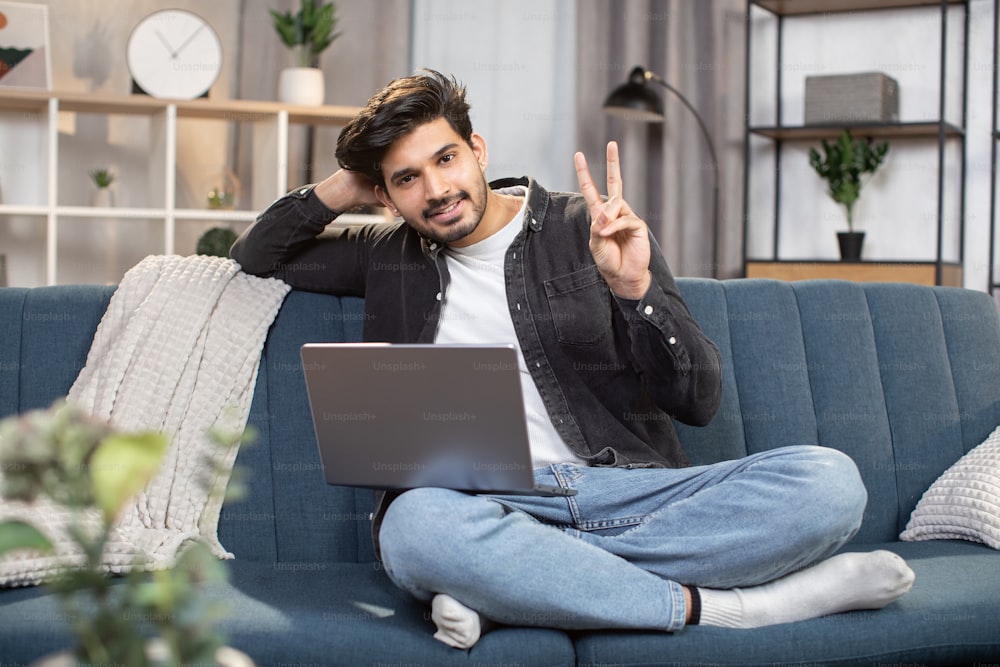 Concepto de jóvenes que usan dispositivos móviles en casa. Hombre indio árabe barbudo sonriente, trabajando en una computadora portátil en casa mientras está sentado en el sofá, mostrando el signo de la victoria a la cámara.
