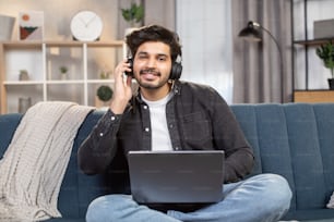 젊은 잘 생긴 아랍 또는 인도 남자, 집에서 소파에 앉아 노트북 PC에서 작업하고 동시에 이어폰을 사용하여 좋아하는 음악을 듣고.
