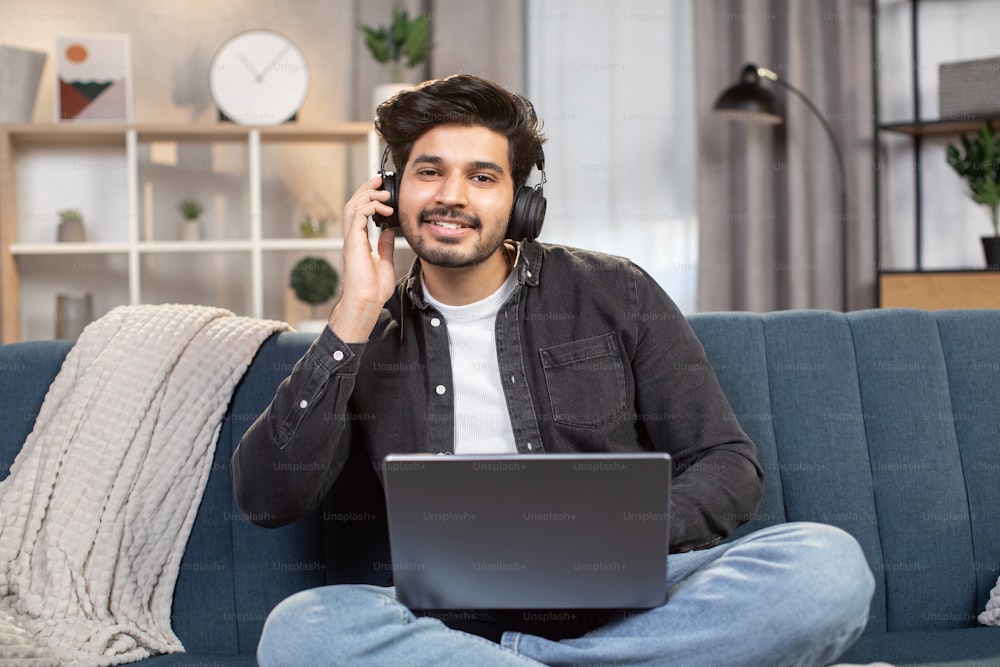 Jovem homem árabe ou indiano bonito, sentado no sofá em casa, trabalhando em laptop pc e ouvindo sua música favorita ao mesmo tempo usando fones de ouvido.