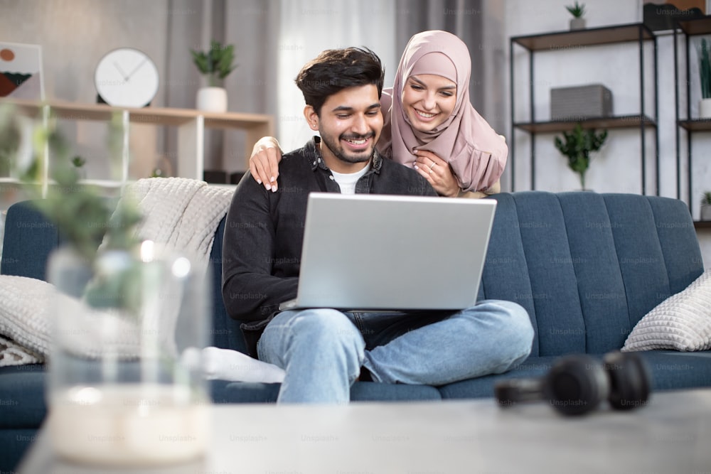 Young handsome bearded arabian man sitting on the sofa and watching movie or news on laptop at home, while his pretty smiling wife in hijab, standing behind him and looking at the screen.
