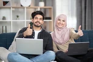 Young multiracial Muslim couple relaxing on sofa at home using laptops, smiling at camera and showing thumbs up.