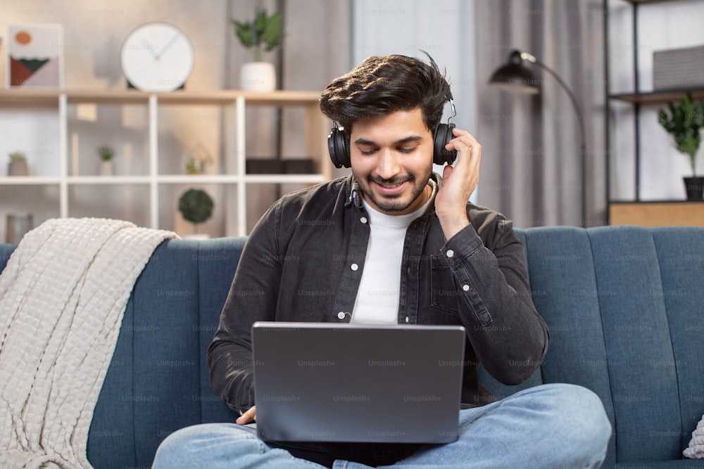 Online lessons, e-learning concept. Young likable Indian man sitting on couch in living room at home, enjoying studying using laptop and headset, looking at screen and listening audio lessons.