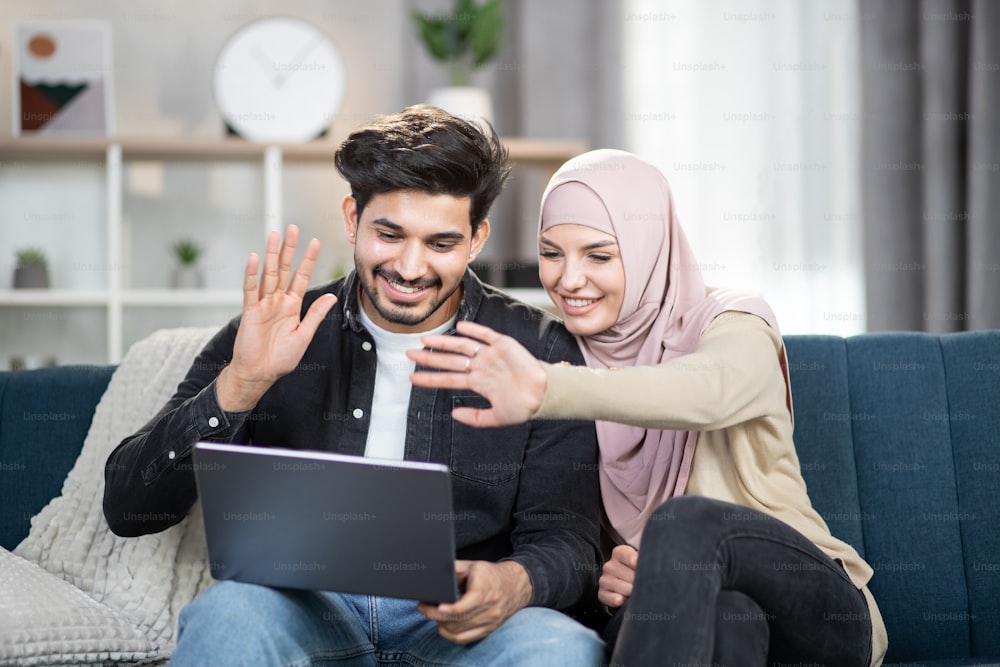 Laughing cheerful young muslim couple sitting on a couch with a laptop at home, waving while having video call talking with relatives or friends. Family, technology, wifi internet concept.