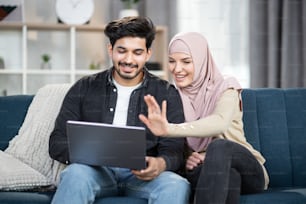 Couple musulman souriant et heureux avec un ordinateur portable à la maison, agitant les mains tout en ayant un appel vidéo avec leurs amis ou leur famille. Concept de rester à la maison pendant la pandémie.