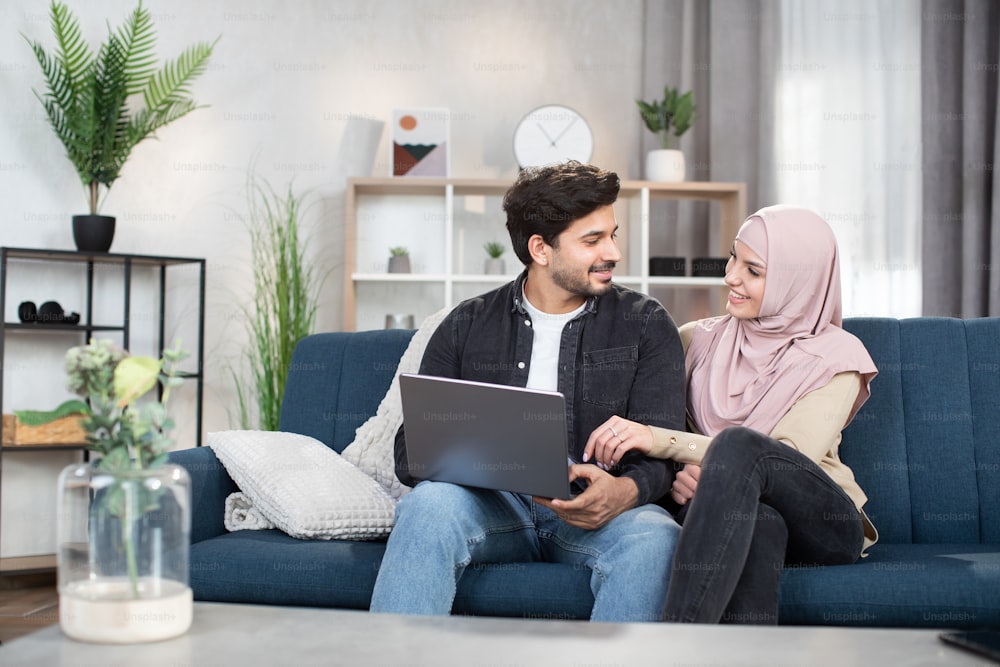 Relaxed Arabian Muslim couple in casual wear, using a laptop pc together for watching online content or shopping, sitting on a sofa in the living room at home.