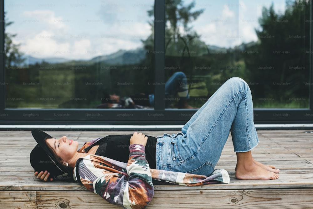 Stylish happy woman relaxing on wooden terrace on background of modern cabin with windows. Young female in casual cloth and hat lying and smiling on porch in mountains. Travel and wanderlust