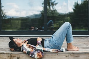 Femme heureuse et élégante se relaxant sur une terrasse en bois sur fond de cabine moderne avec fenêtres. Jeune femme en tissu décontracté et chapeau couchée et souriante sur le porche dans les montagnes. Voyage et envie de voyager