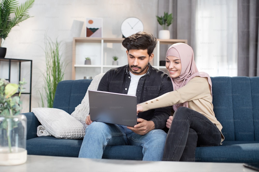 Family leisure, technology concept. Lovely happy young arabic muslim couple in casual domestic clothes, sitting together on soft couch at home and watching interesting movie on laptop.