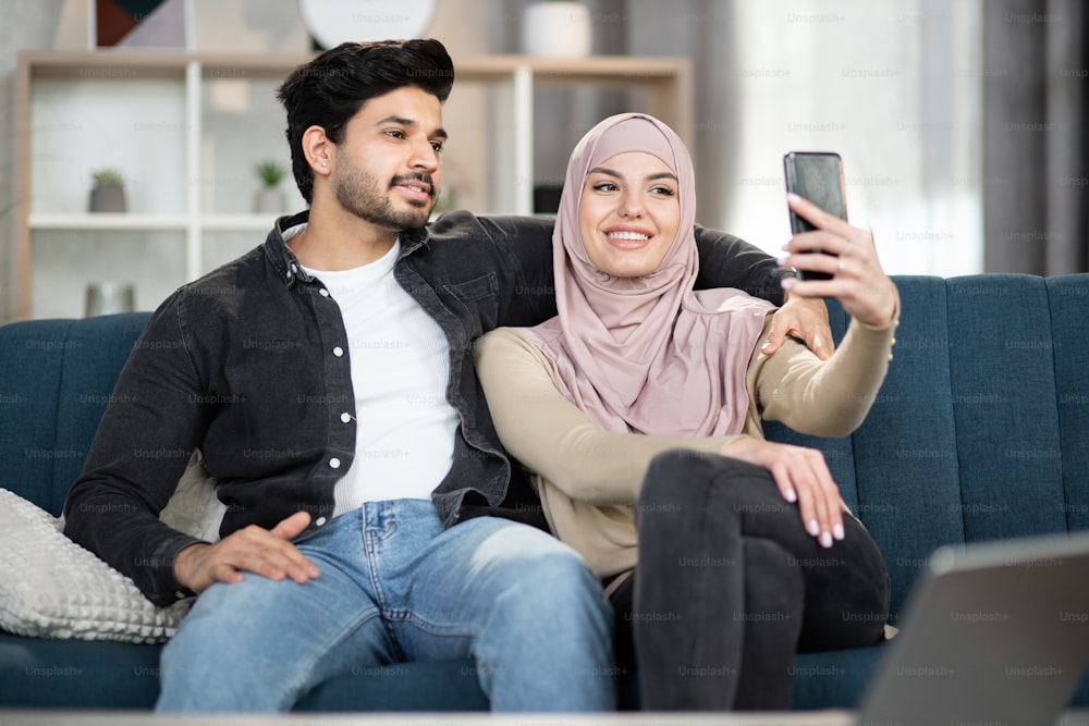 Vue rapprochée de face d’un jeune couple musulman charmant et joyeux assis sur un canapé à la maison, utilisant un téléphone portable et faisant une photo selfie ensemble.