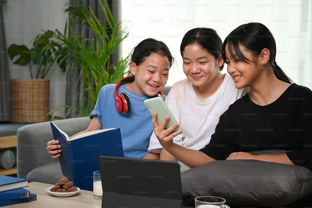 세 명의 아시아 소녀가 주말에 스마트폰을 사용하고 집에서 소파에 앉아 즐거운 시간을 보내고 있다.
