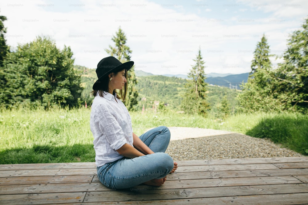 Stylish hipster woman relaxing on wooden terrace on background of sunny mountains hills. Happy young female in hat and casual cloth sitting on porch. Calm tranquil moment. Travel and wanderlust