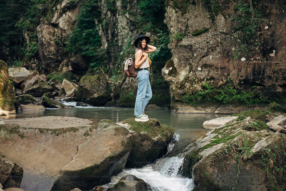 배낭을 메고 산에서 강을 배경으로 휴식을 취하는 세련된 여성 여행자. 캐주얼한 천과 모자를 쓴 젊은 여성이 강가의 바위 위에 서서 웃고 있다. 여행과 방랑벽. 숲 탐험