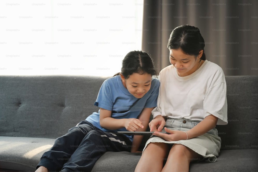 Asiatisches Mädchen und ihre Schwester benutzen ein digitales Tablet zusammen, während sie auf dem Sofa im Wohnzimmer sitzen.