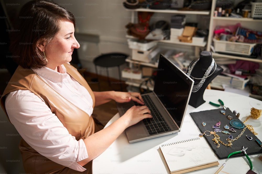 Side view of a smiling female jewelry designer sitting at the laptop in her workshop