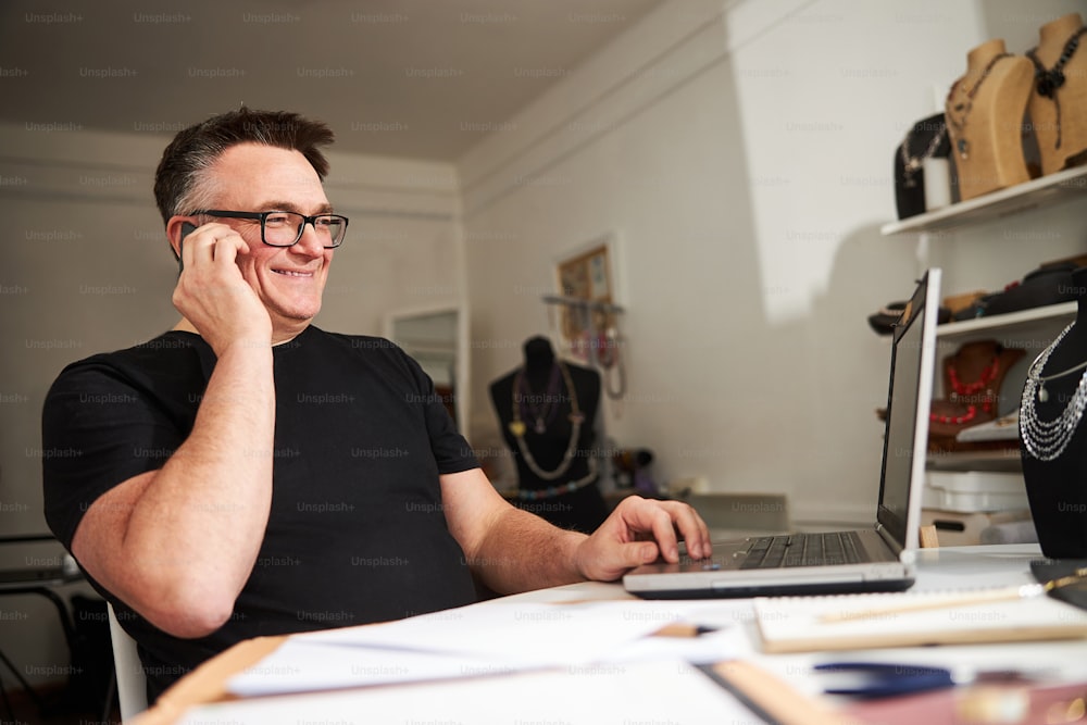 Lächelnder zufriedener Mann mit Brille sitzt während des Telefongesprächs am Laptop in seiner Werkstatt
