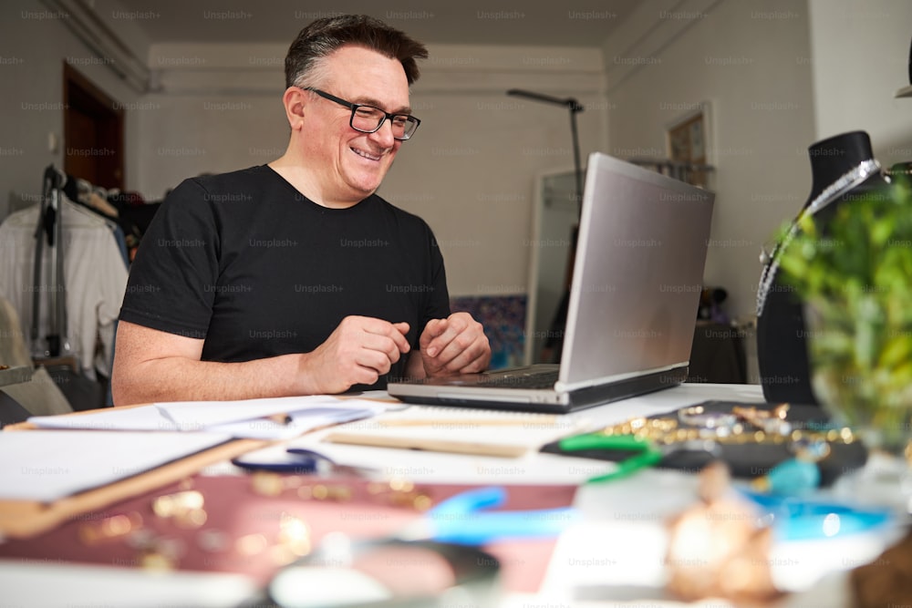 Portrait of a professional joyful male jewelry designer sitting at the laptop in his workshop